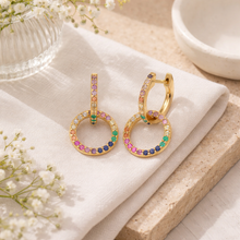 Load image into Gallery viewer, Gold hoop earrings with colorful gemstones on a white cloth with flowers and a bowl in the background.
