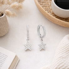 Load image into Gallery viewer, Star-shaped silver earrings on a light surface with a cup and dried plants in the background.
